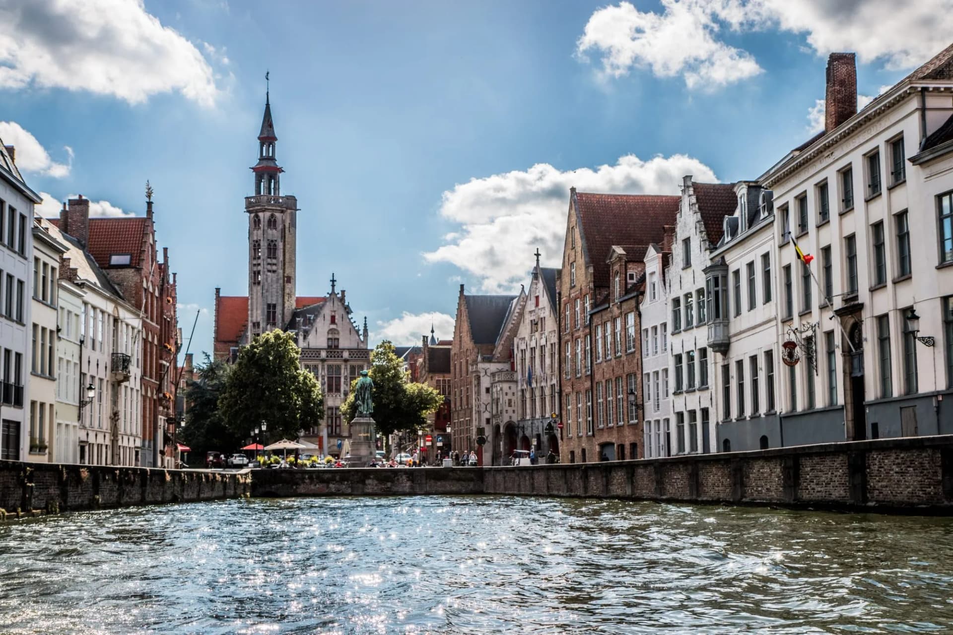 Canal with sunlit water reflecting light, flanked by historic buildings and a tall spire under a blue sky.