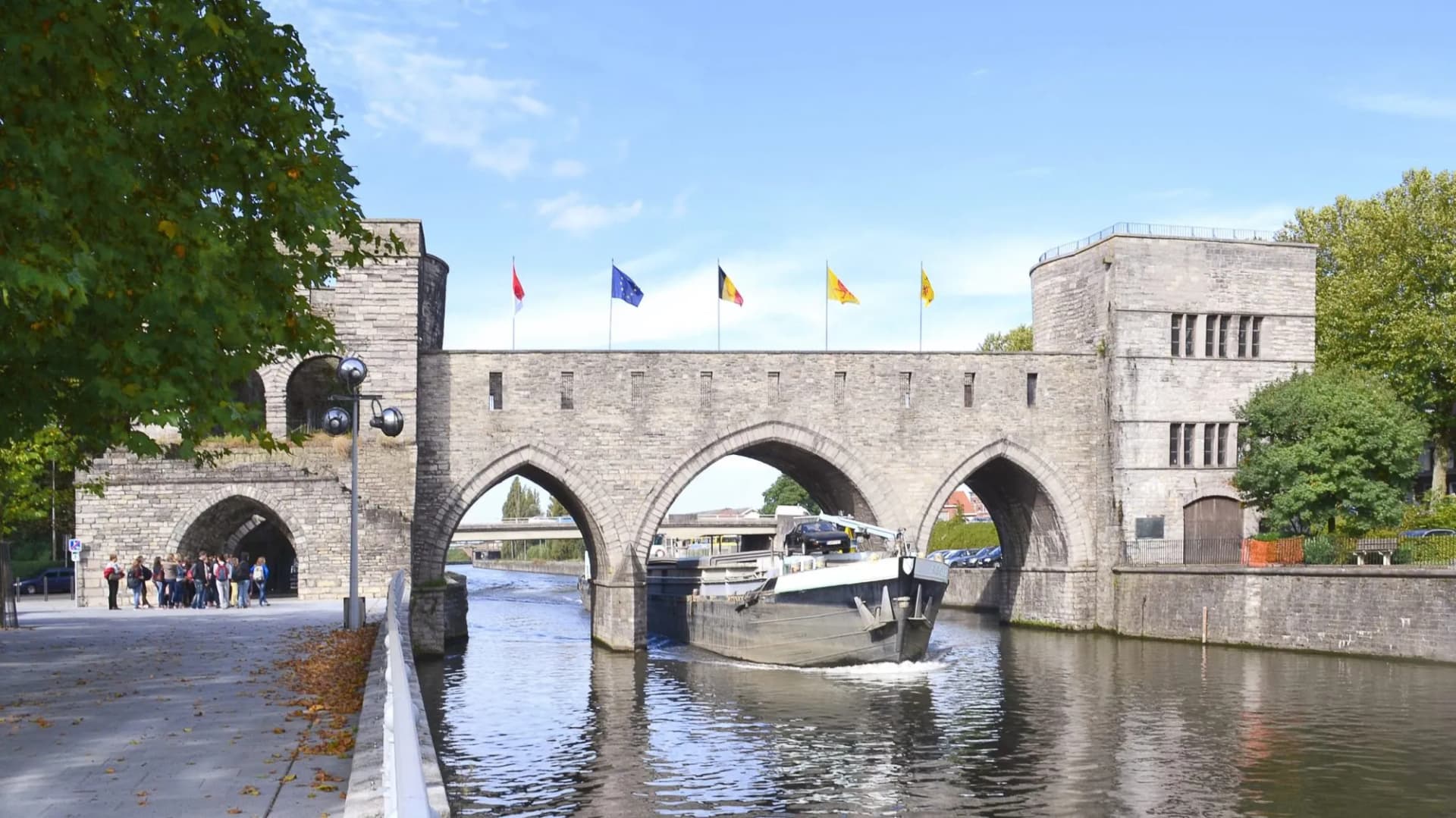 Cargo boat passes under stone bridge with flags; group of people on riverside walkway.