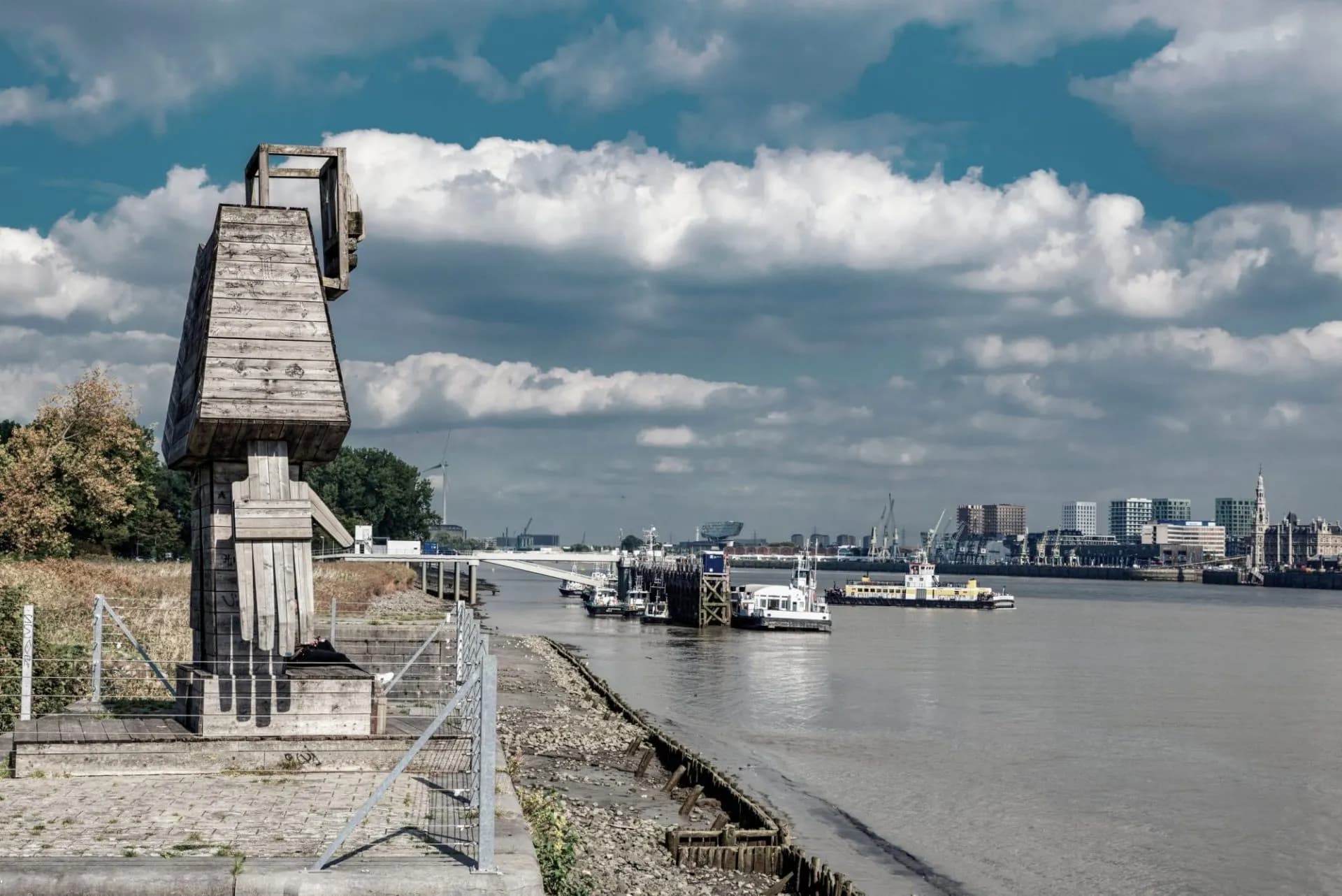 Wooden sculpture by the river with boats and Antwerp skyline under cloudy sky