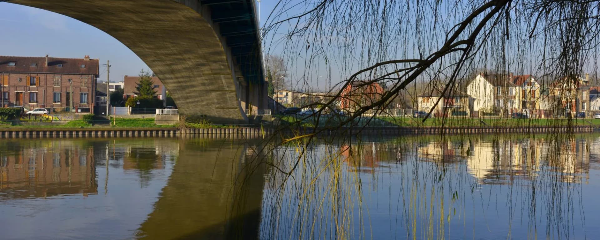 Concrete bridge arch over river with weeping willow branches and houses in Conflans-Sainte-Honorine.