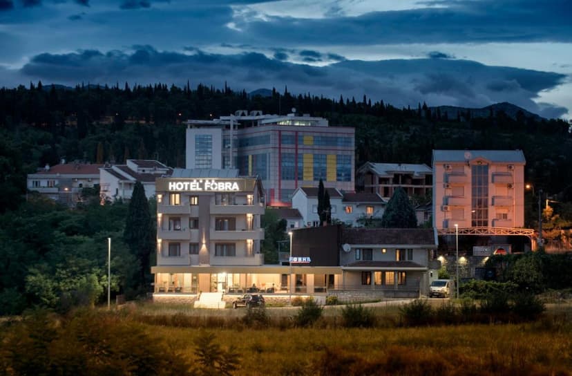 Hotel Fobra Podgorica illuminated at dusk with dark, forested hills in the background.