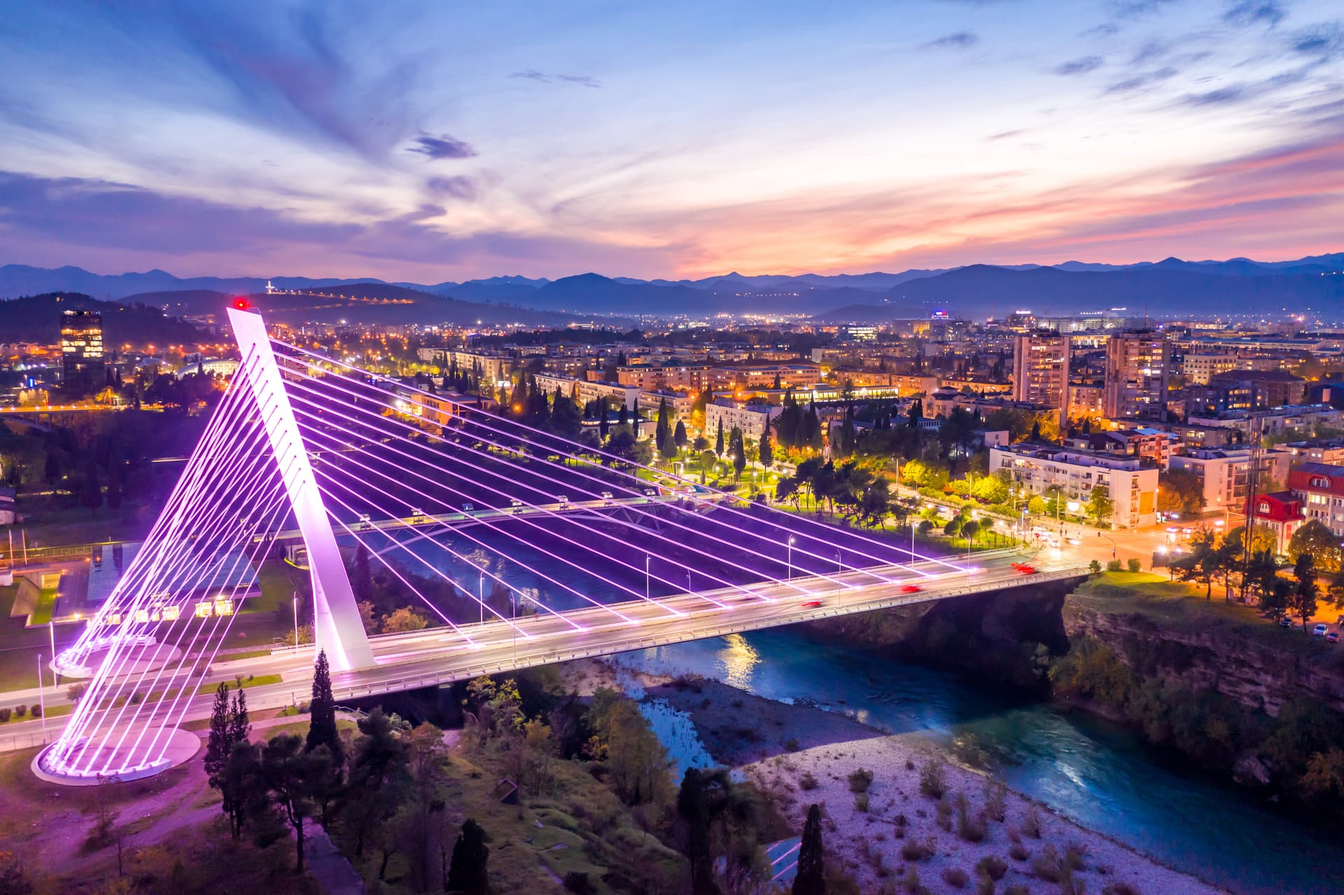 Millennium Bridge illuminated purple over river at dusk with Podgorica city lights and mountains in background.