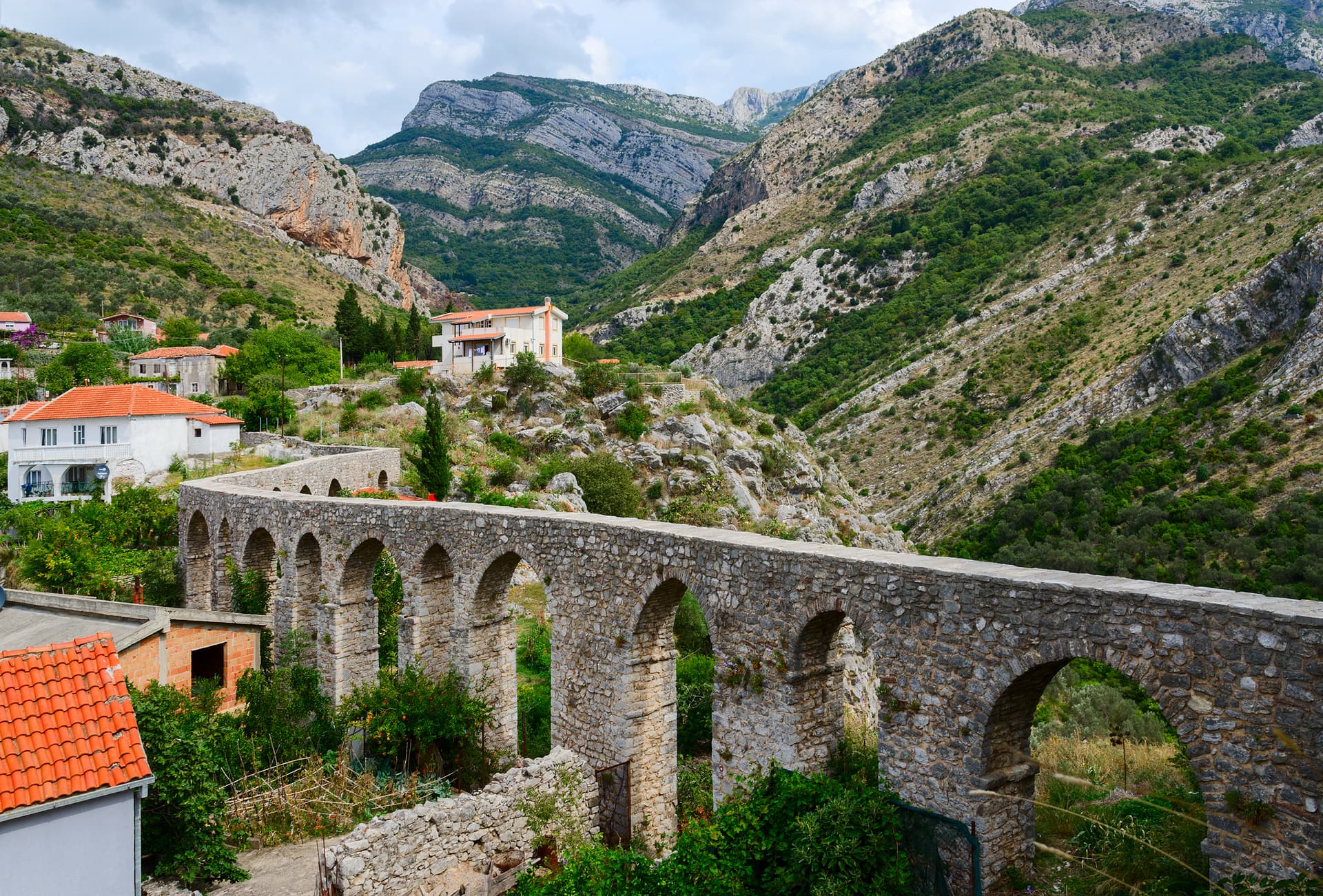 Stone aqueduct arches in a valley with houses and steep, green mountains in Bar, Montenegro.