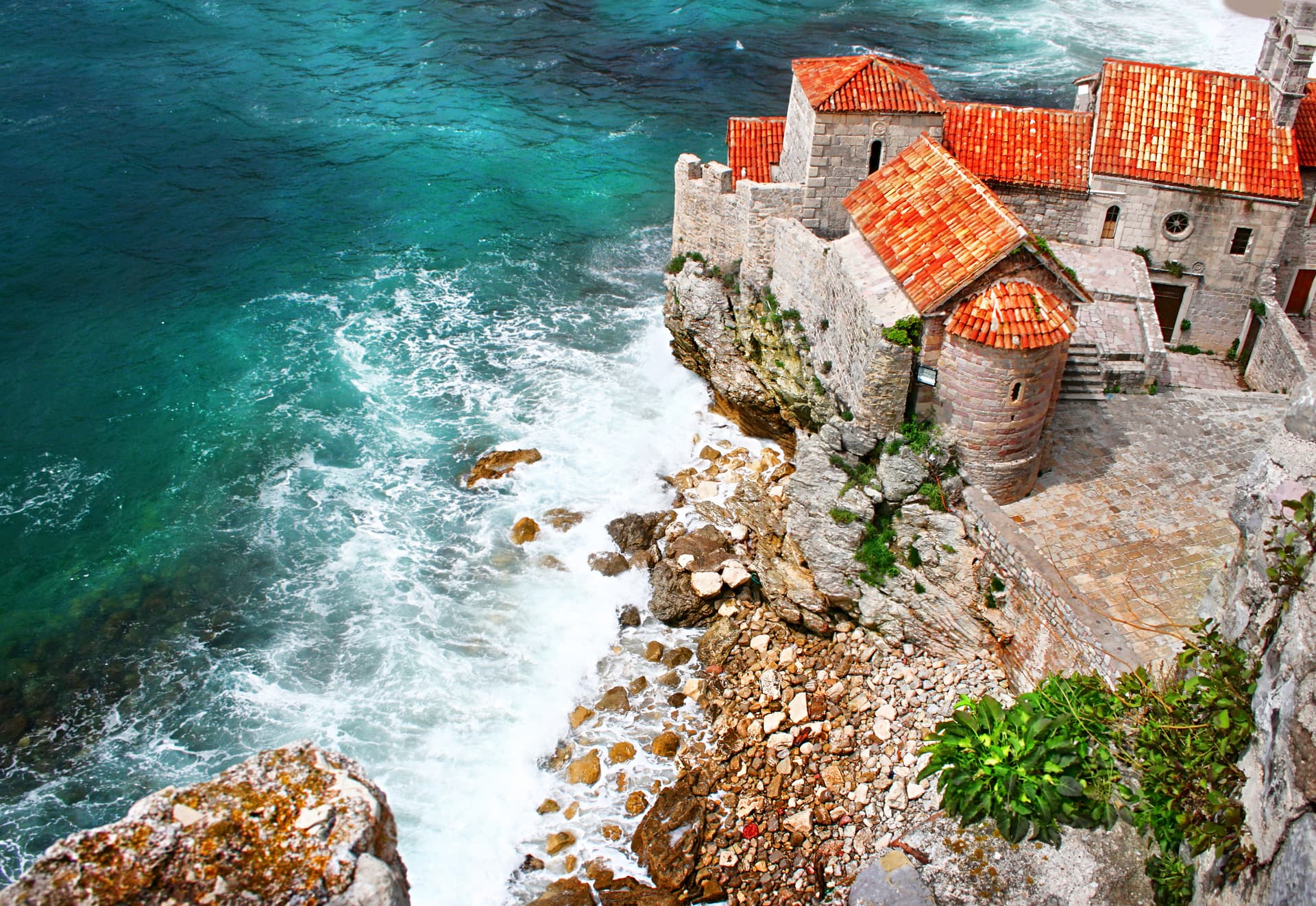 Stone buildings with red tile roofs on a cliff above turquoise sea waves in Old Town Budva, Montenegro.