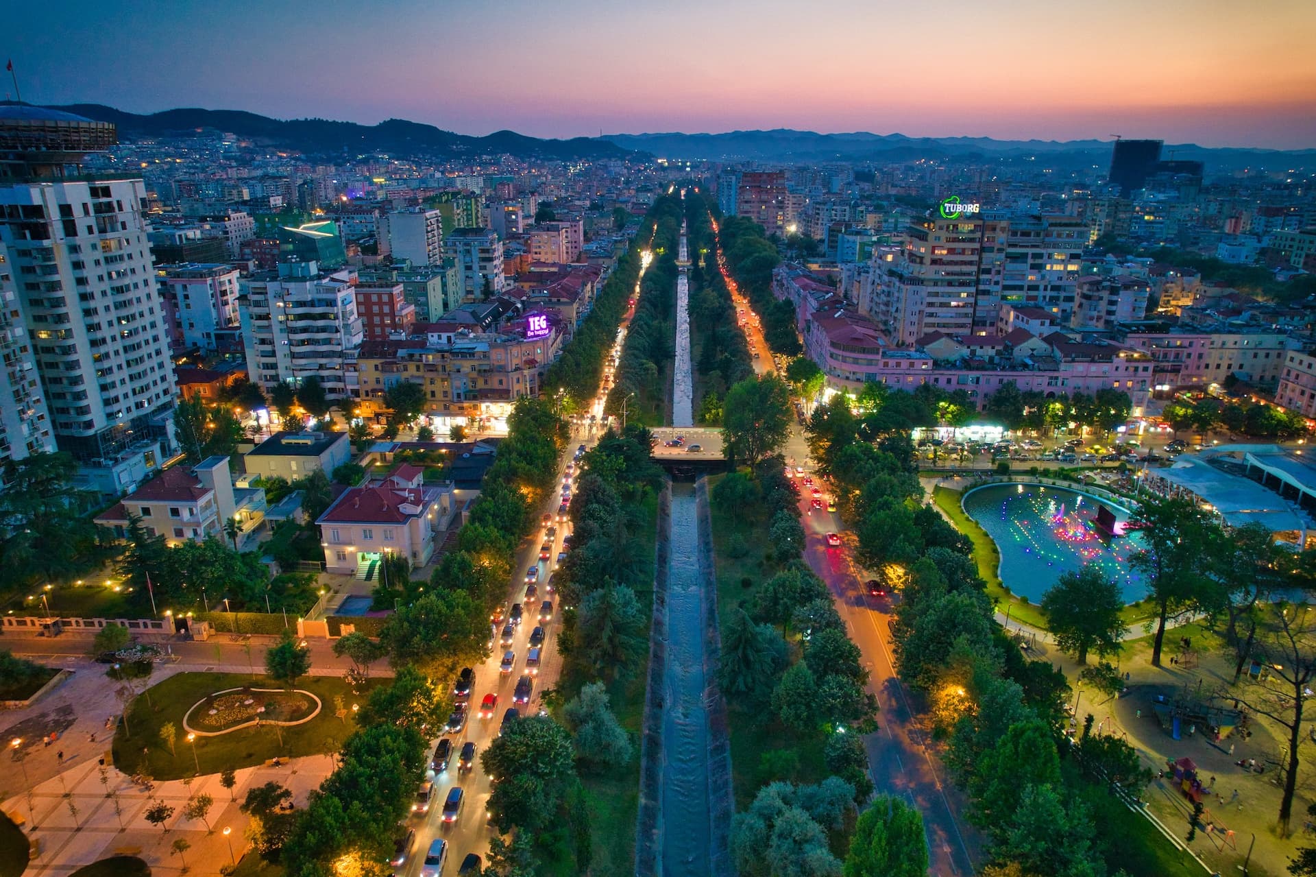 Aerial view of Tirana, Albania at night with a central canal, tree-lined roads, and illuminated city buildings.