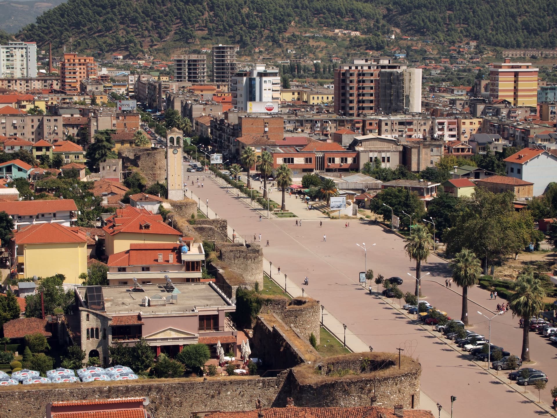 Cityscape of Elbasan, Albania, featuring a central square, clock tower, and buildings against a steep, forested hill.