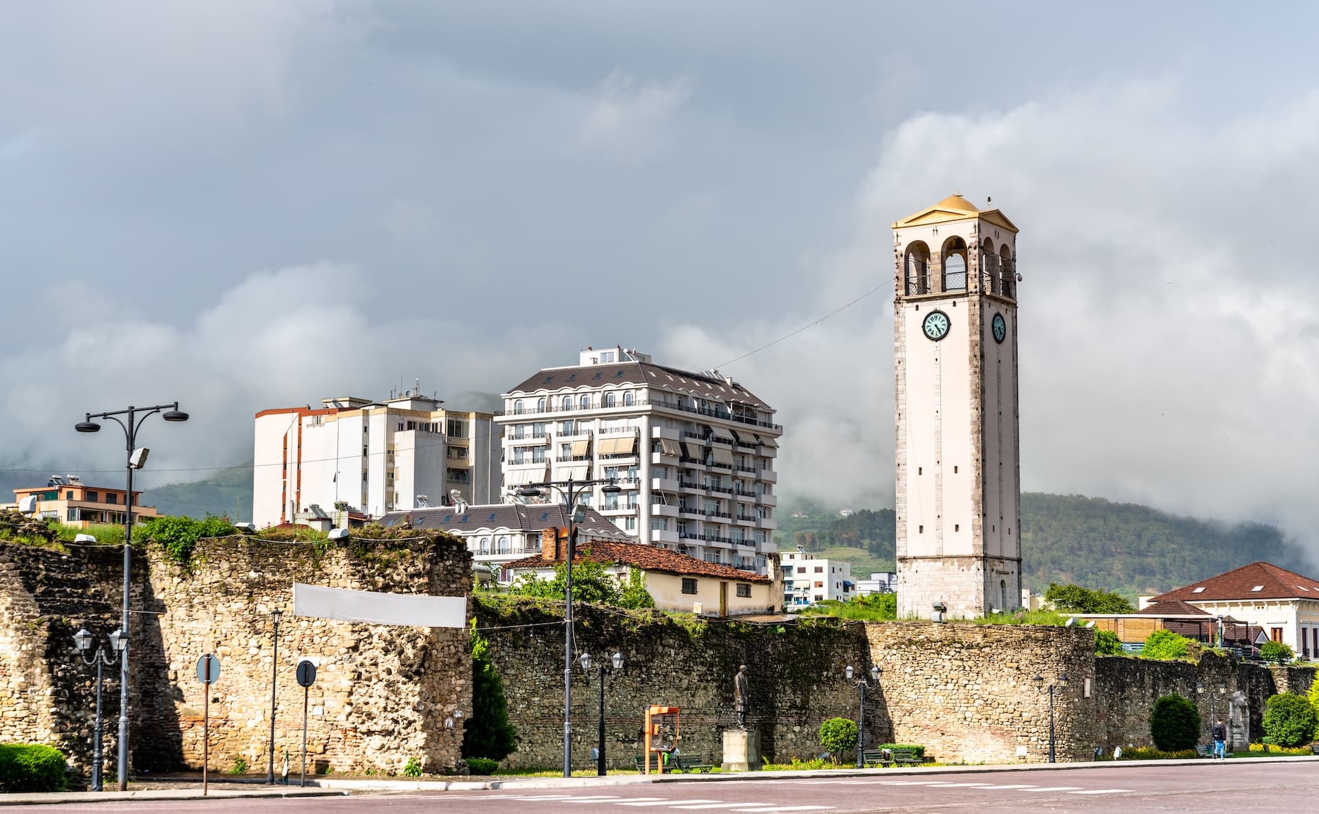 Clock tower above stone fortress walls with modern buildings and green hills in Elbasan, Albania.