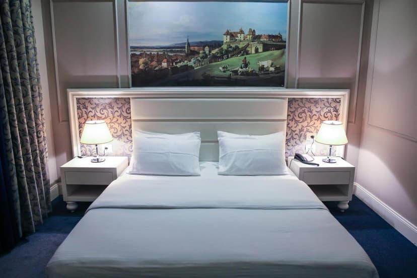 Hotel Monarch Elbasan room with white bedding, bedside lamps, and a painting above the headboard.