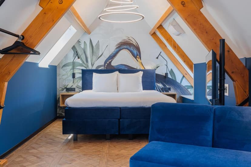 Attic hotel room with blue velvet bed, tropical bird mural, and exposed wood beams.