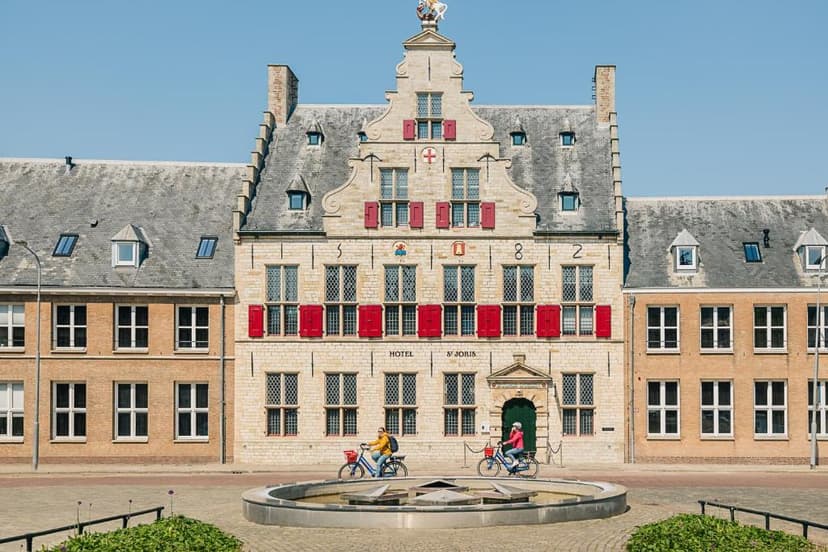 Two people cycling past Hotel St. Joris with stepped gable and red shutters in a town square.