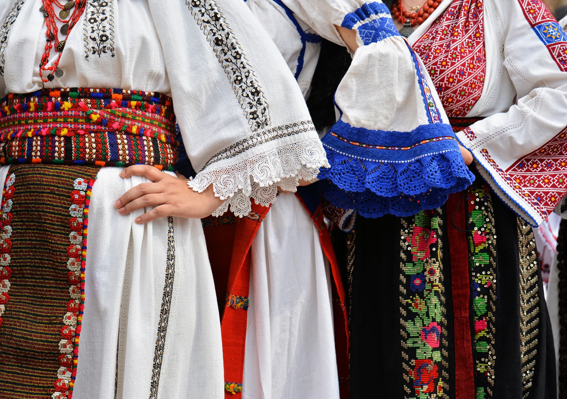 Close-up of traditional Romanian Dobrogea costumes with white embroidery, colorful woven belts, and red beads.
