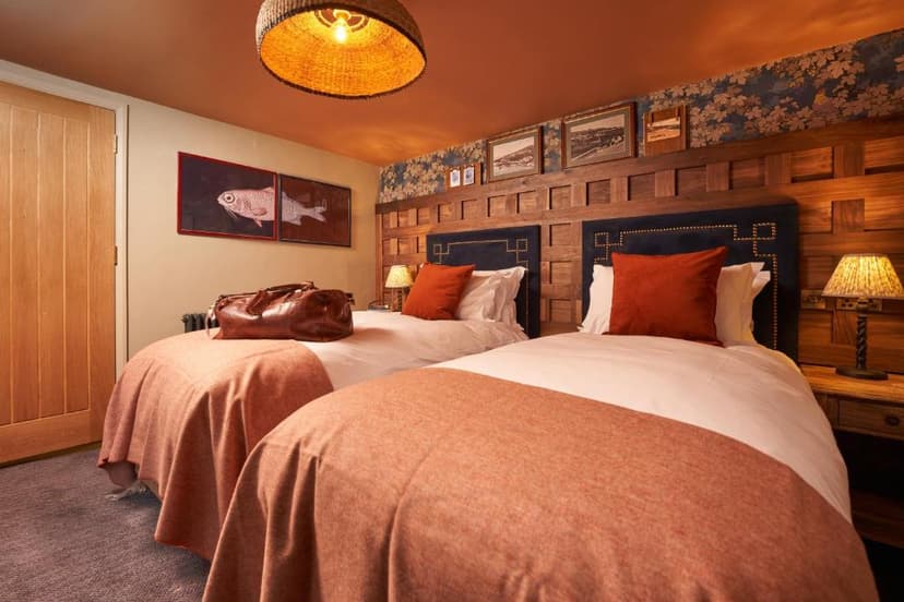 Twin hotel beds with white linens, orange throws, and a leather travel bag in a warmly lit room.