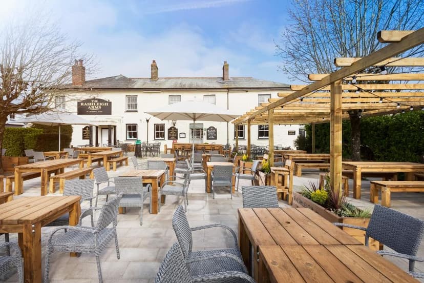 Outdoor patio dining area with wooden tables, grey chairs, and The Rashleigh Arms pub facade.