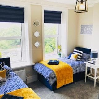 Twin bedroom with blue and yellow bedding, white side table, and view of green trees outside windows.