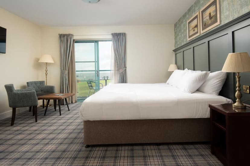 Hotel bedroom with plaid carpet, white bedding, and balcony view of green field.