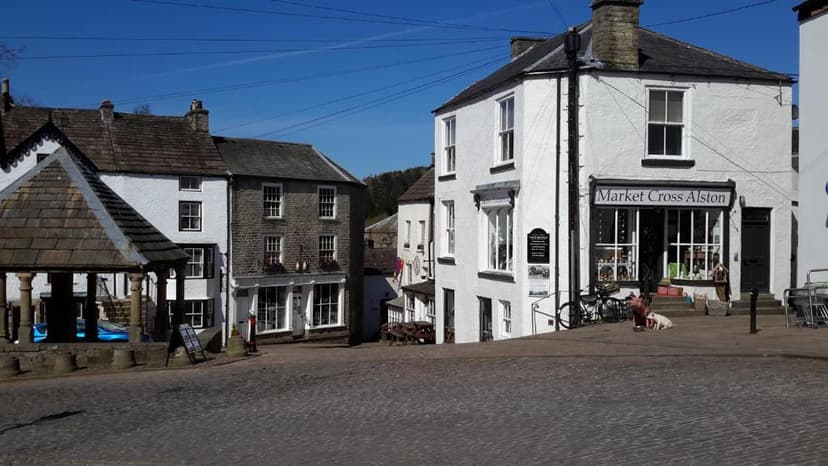 Cobblestone square with Market Cross Alston shop and stone market shelter under blue sky.