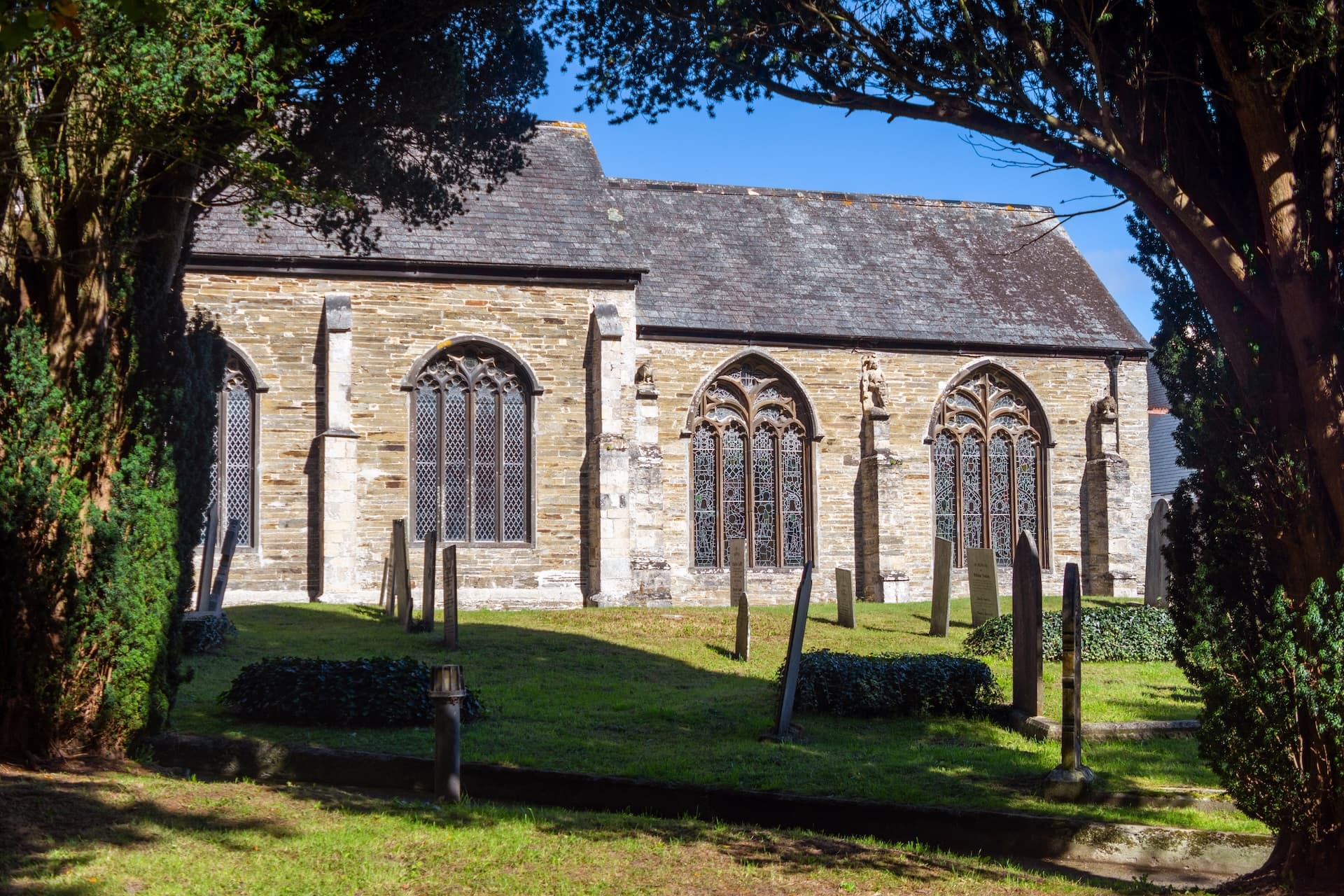 Stone church exterior with stained glass windows viewed through dark trees in a churchyard.