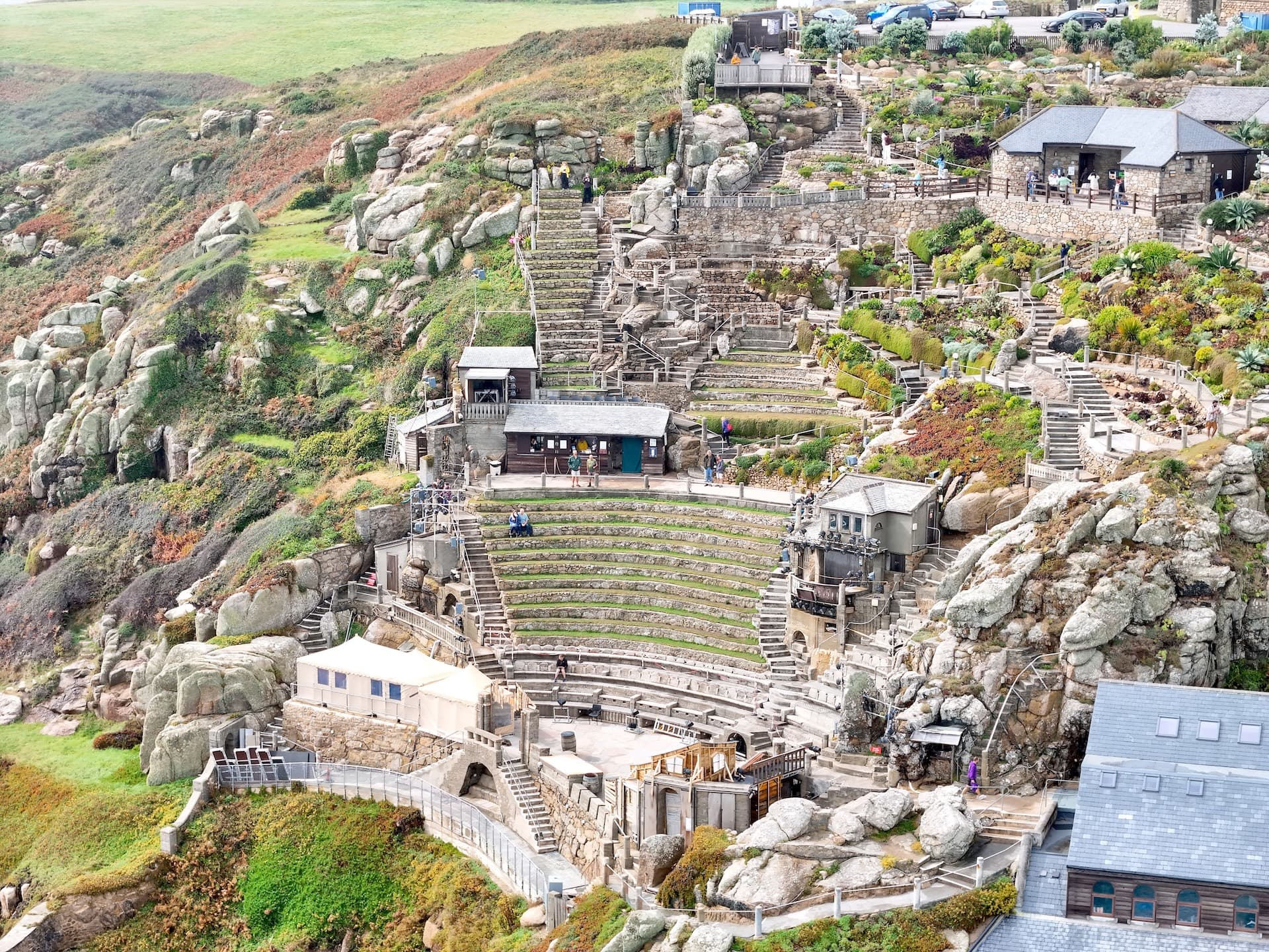 Outdoor amphitheater built into a rocky, green hillside with stone seating tiers and small buildings.