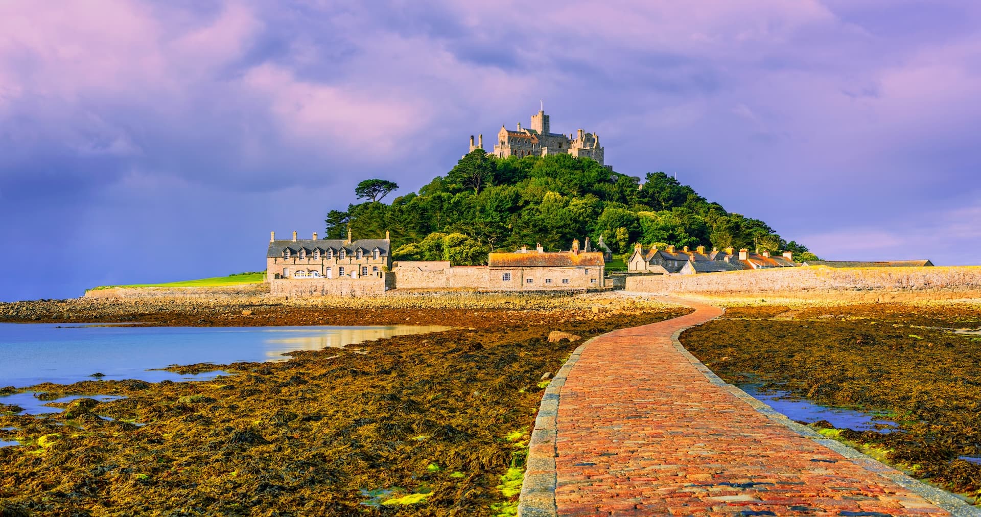 St Michael's Mount castle on wooded hill approached by tidal causeway under dramatic sky.