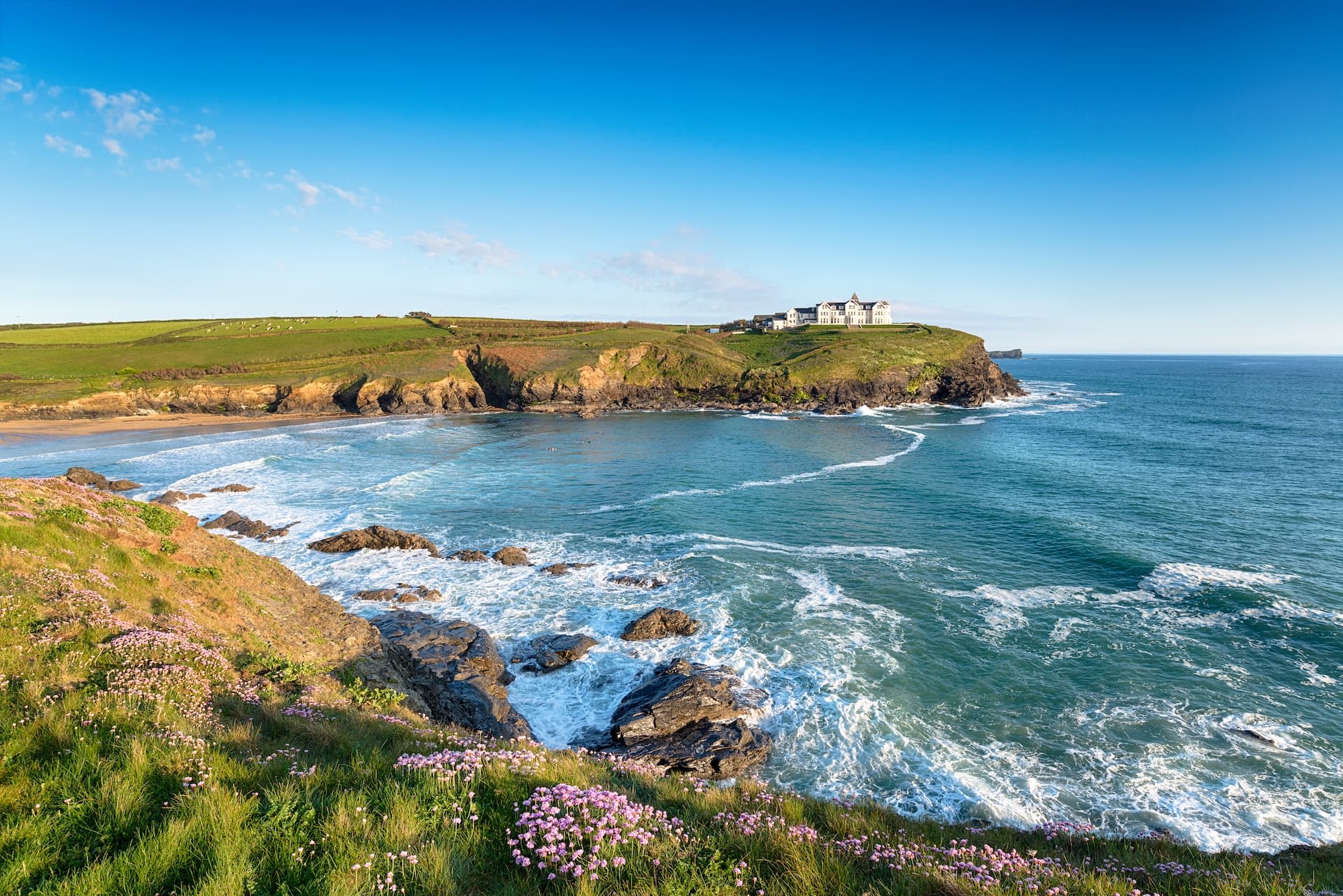 Coastal cliffs with pink sea thrift flowers overlooking turquoise ocean waves and a white hotel.
