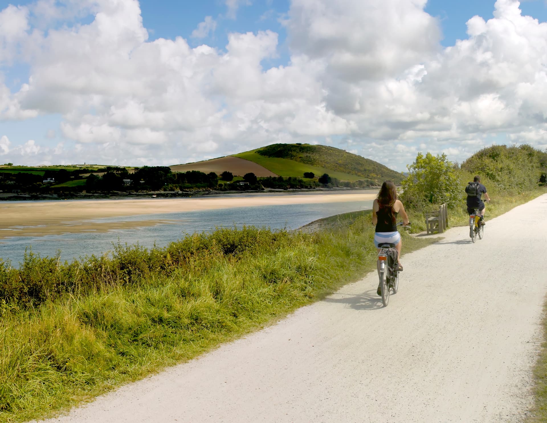 Two people cycling on a dirt path beside a river estuary and green hills