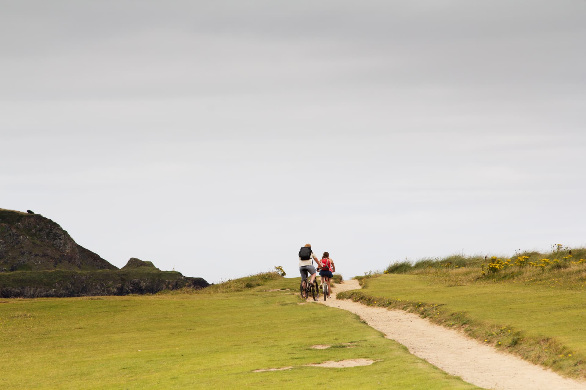 Two people cycling on a dirt path across grassy coastal hills under an overcast sky.