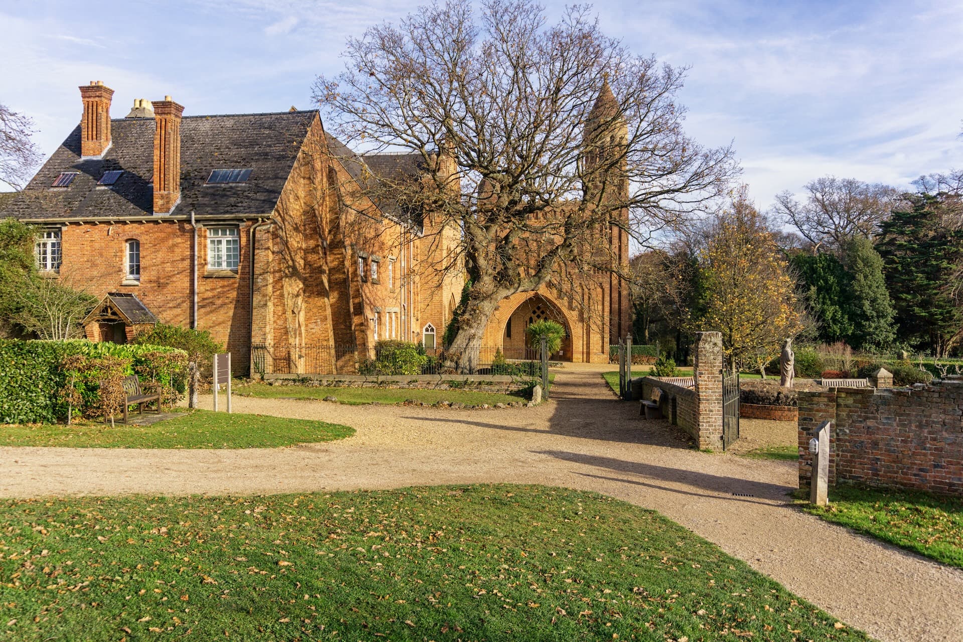 Brick abbey building with large tree in foreground, gravel path, and autumn lawn.