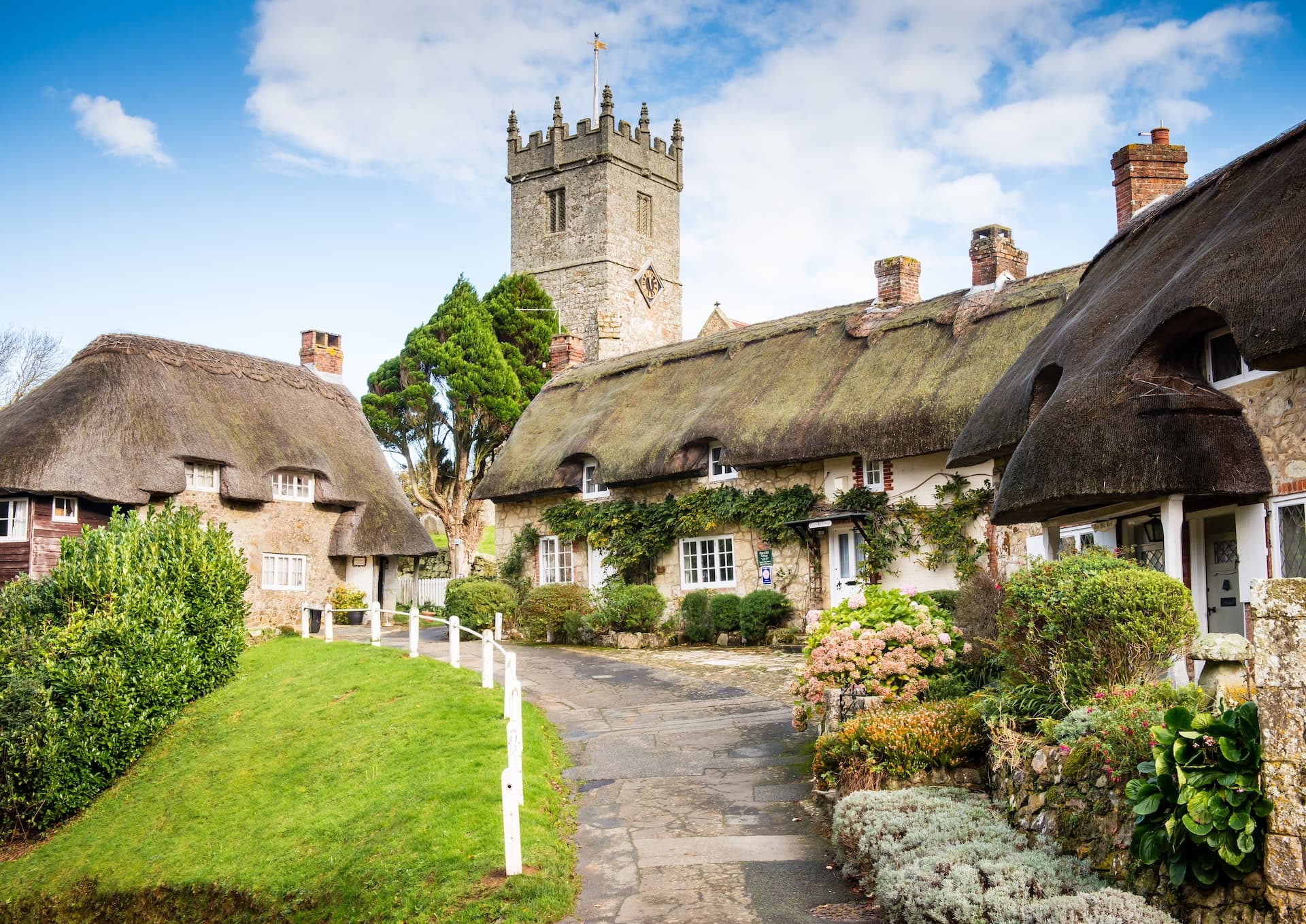 Thatched roof cottages and a stone church tower in a green English village setting.