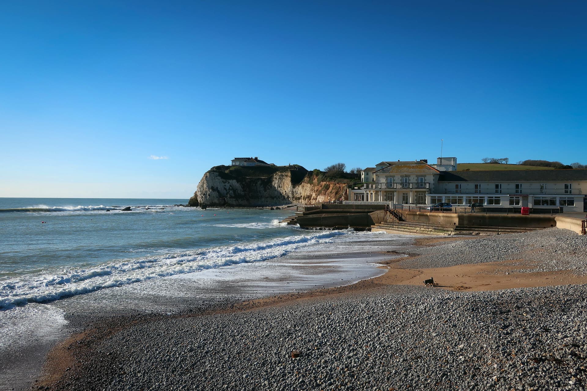 Pebble beach with waves washing ashore near a white cliff and seaside hotel, Freshwater Bay.