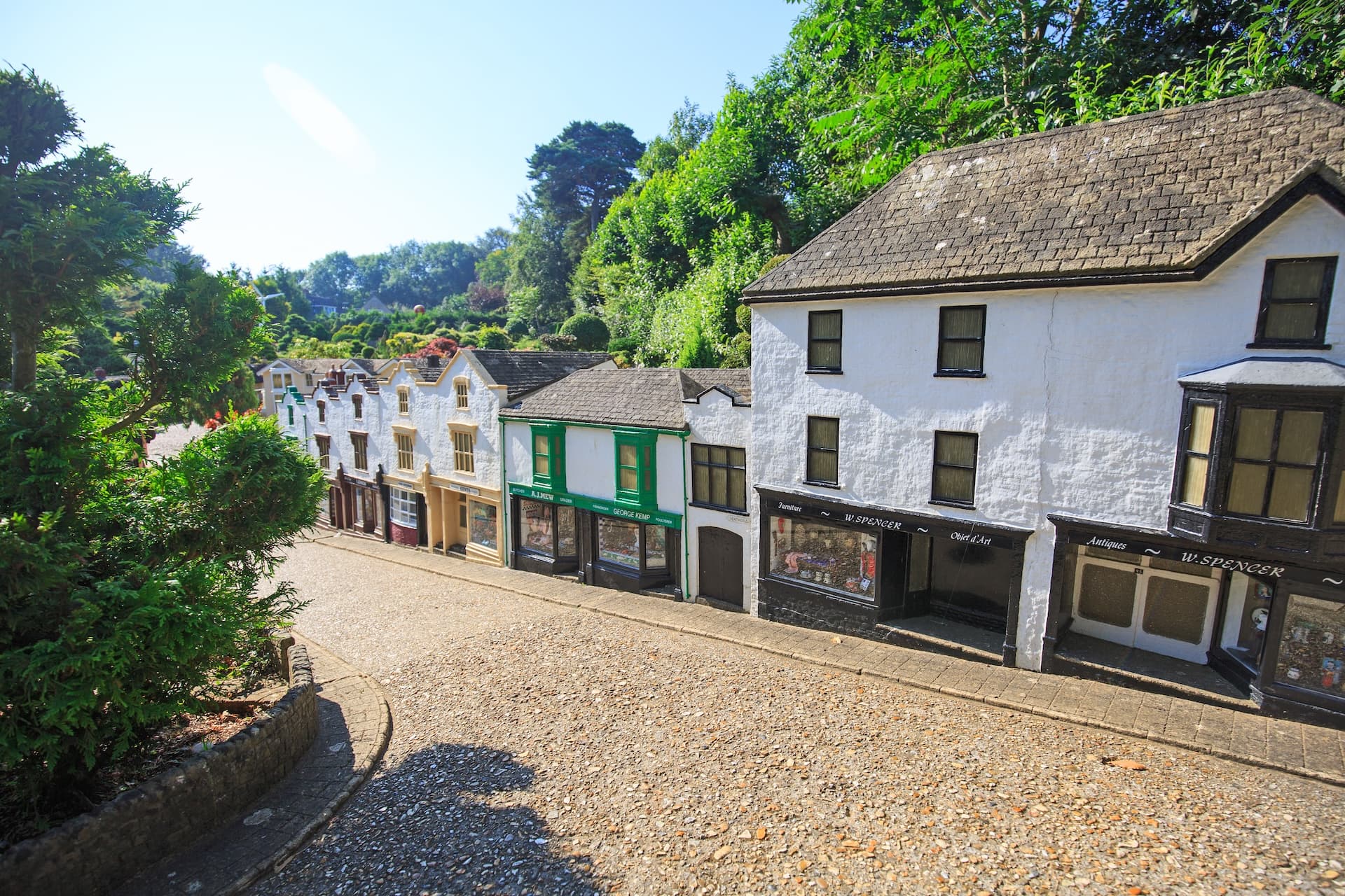 Miniature village street with white shops, cobblestones, and lush green hillside under bright sun.