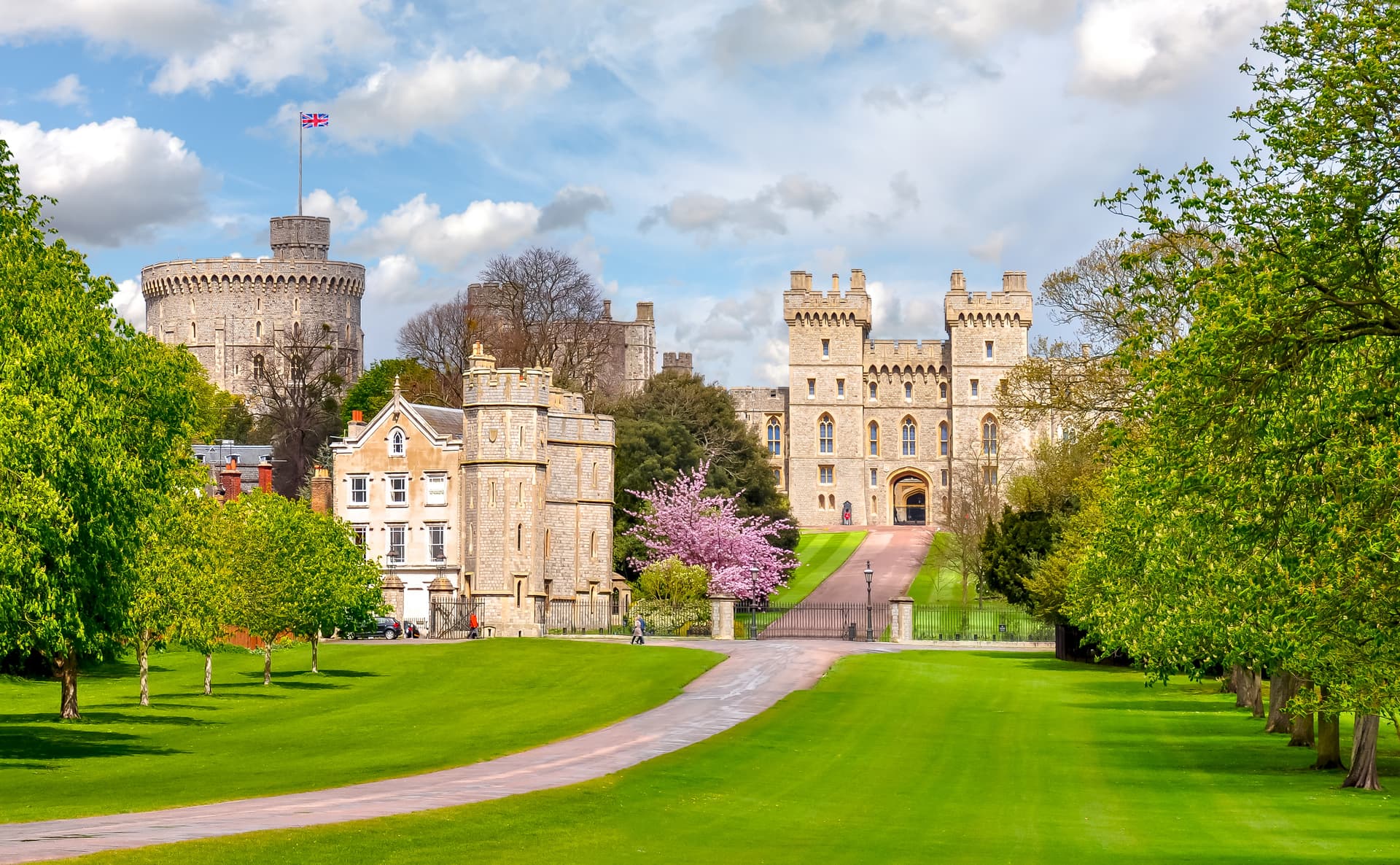 Windsor Castle with Union Jack flying, green lawns, and spring blossoms under a cloudy blue sky.
