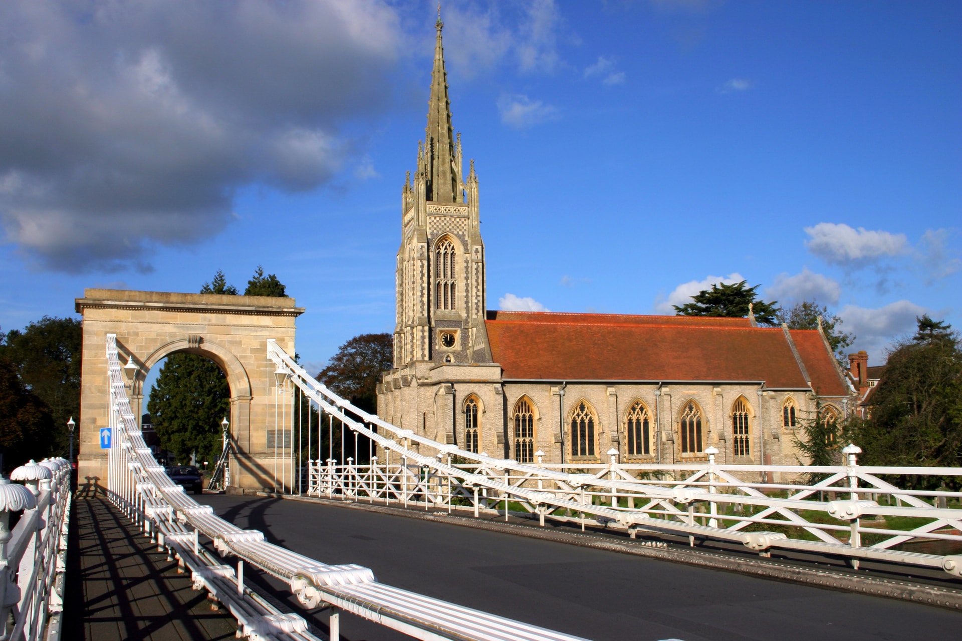 Suspension bridge with white railings leading toward a stone church with a tall spire.