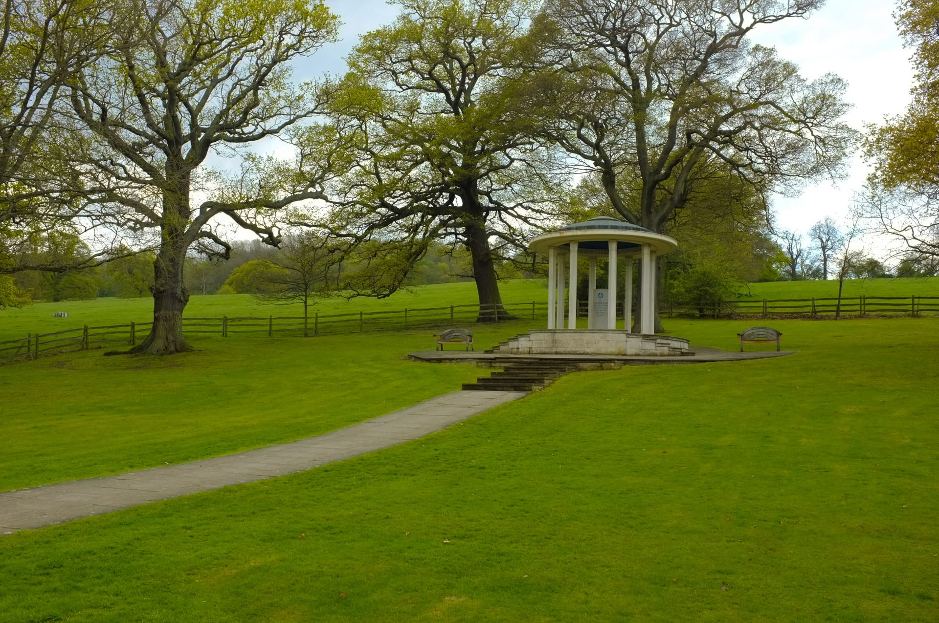 White circular memorial structure with columns on a grassy lawn with large trees.