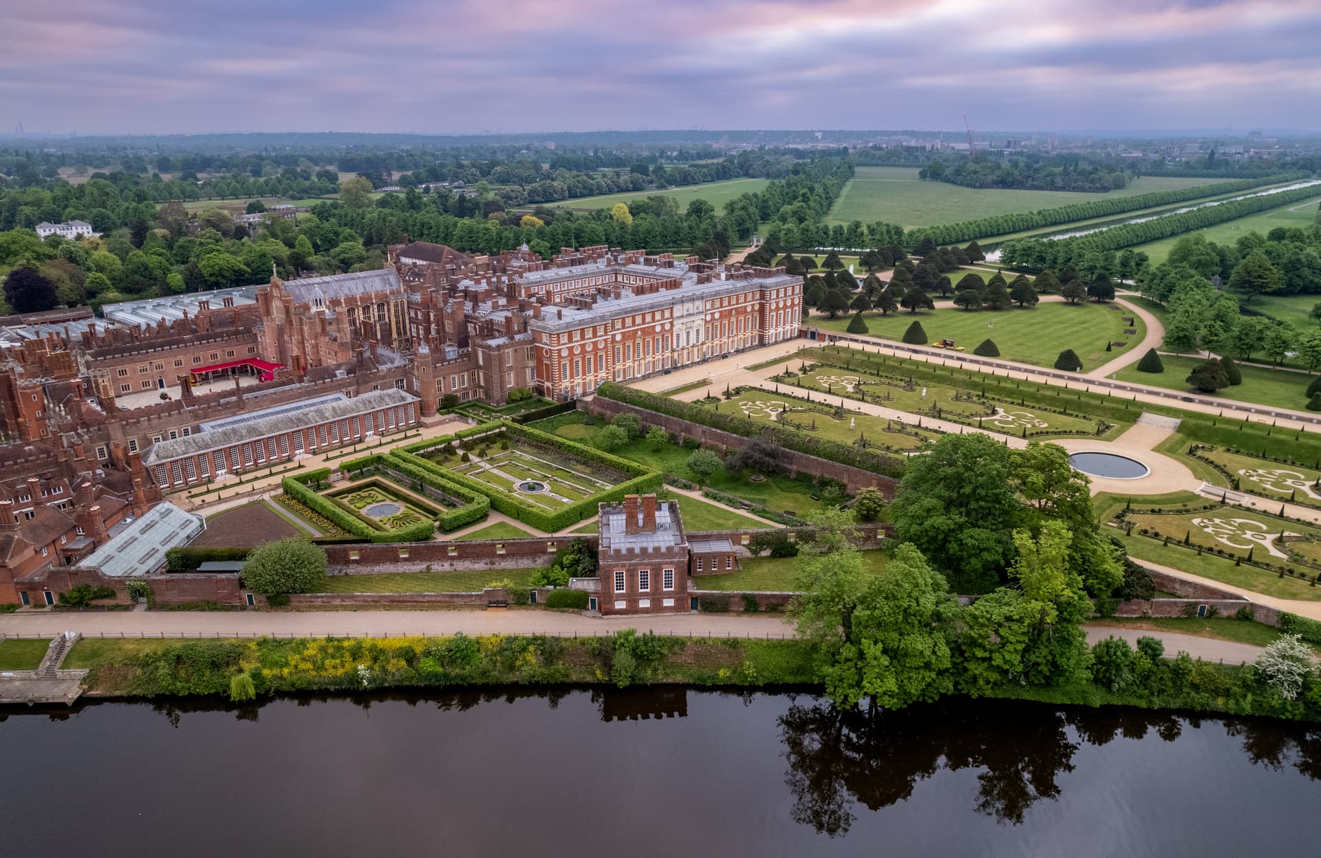 Aerial view of Hampton Court Palace with formal gardens and river in the foreground.