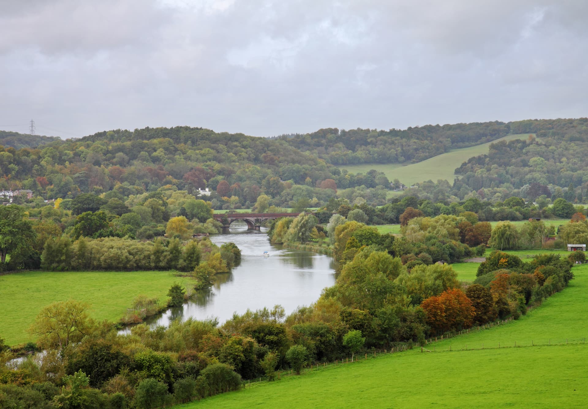 River scene with autumn trees, a boat, and a brick bridge under a cloudy sky.