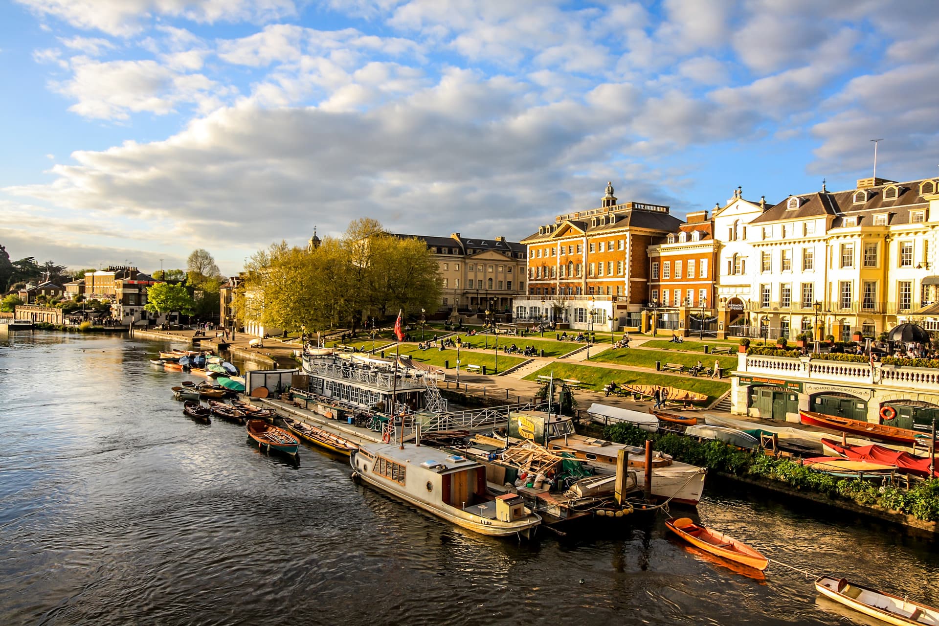 Boats moored on river next to historic buildings and green lawn under cloudy sky.