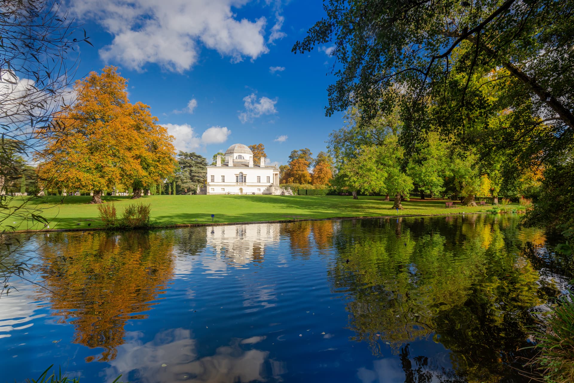 White Palladian villa reflected in water with autumn trees under a blue sky in Chiswick.