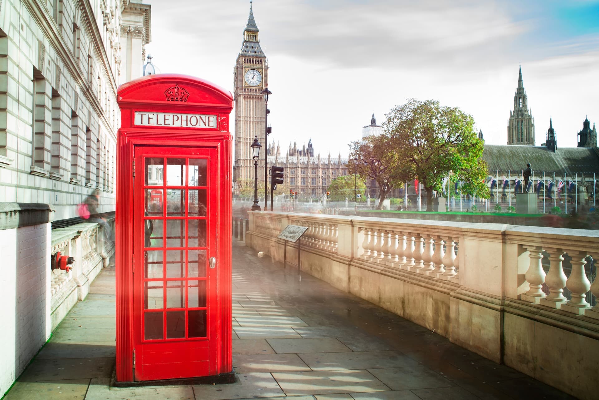 Red telephone box on sidewalk with Big Ben and Houses of Parliament in London background.