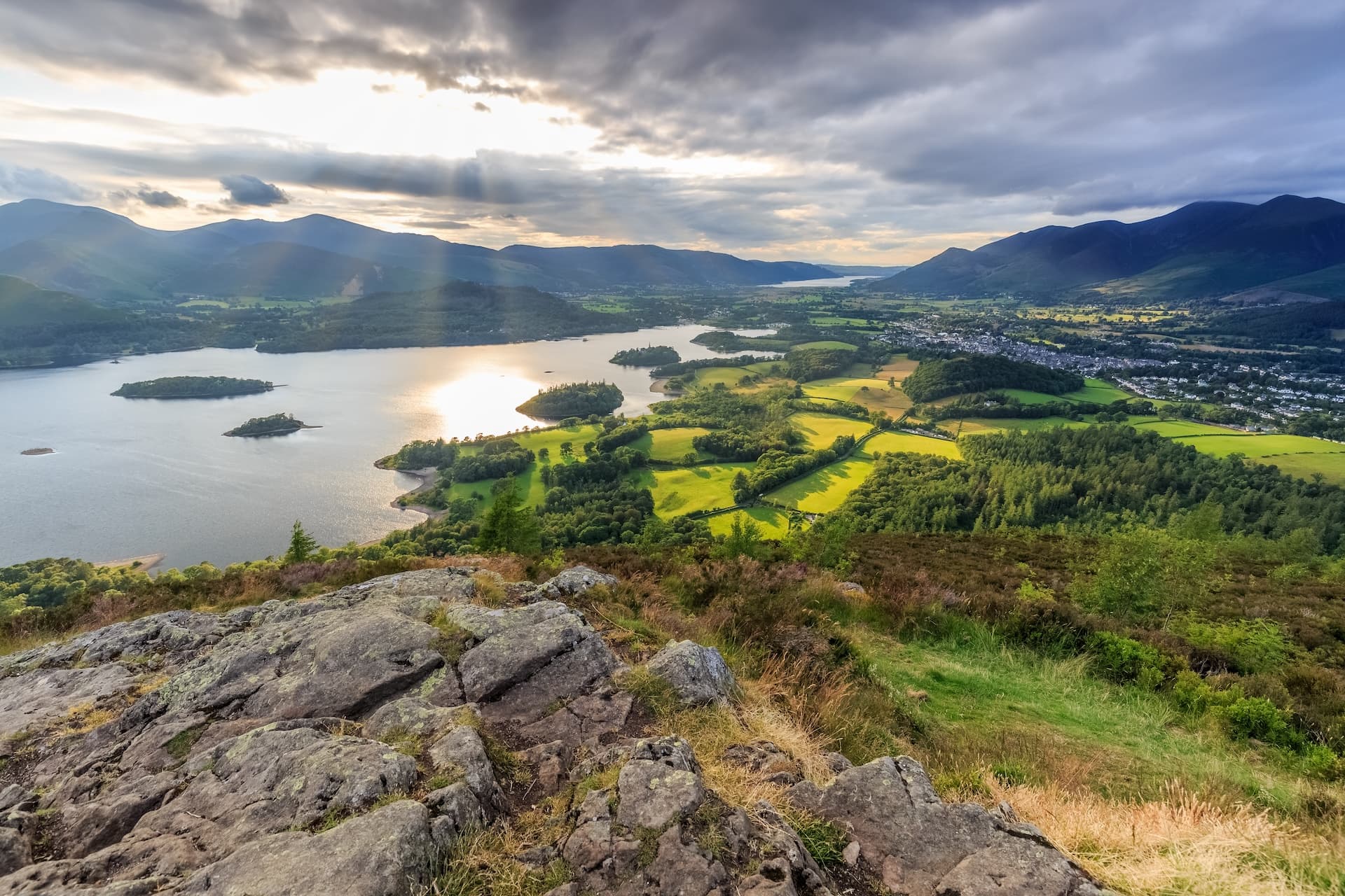 View from rocky outcrop over Derwentwater, Keswick, and surrounding mountains under dramatic sky.