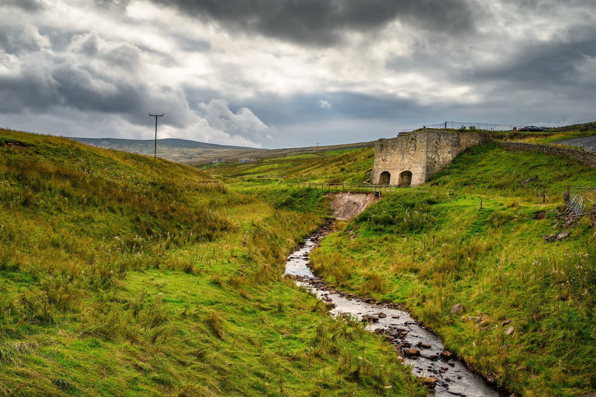 Stone lime kiln structure beside a stream in a green, hilly landscape under a cloudy sky.