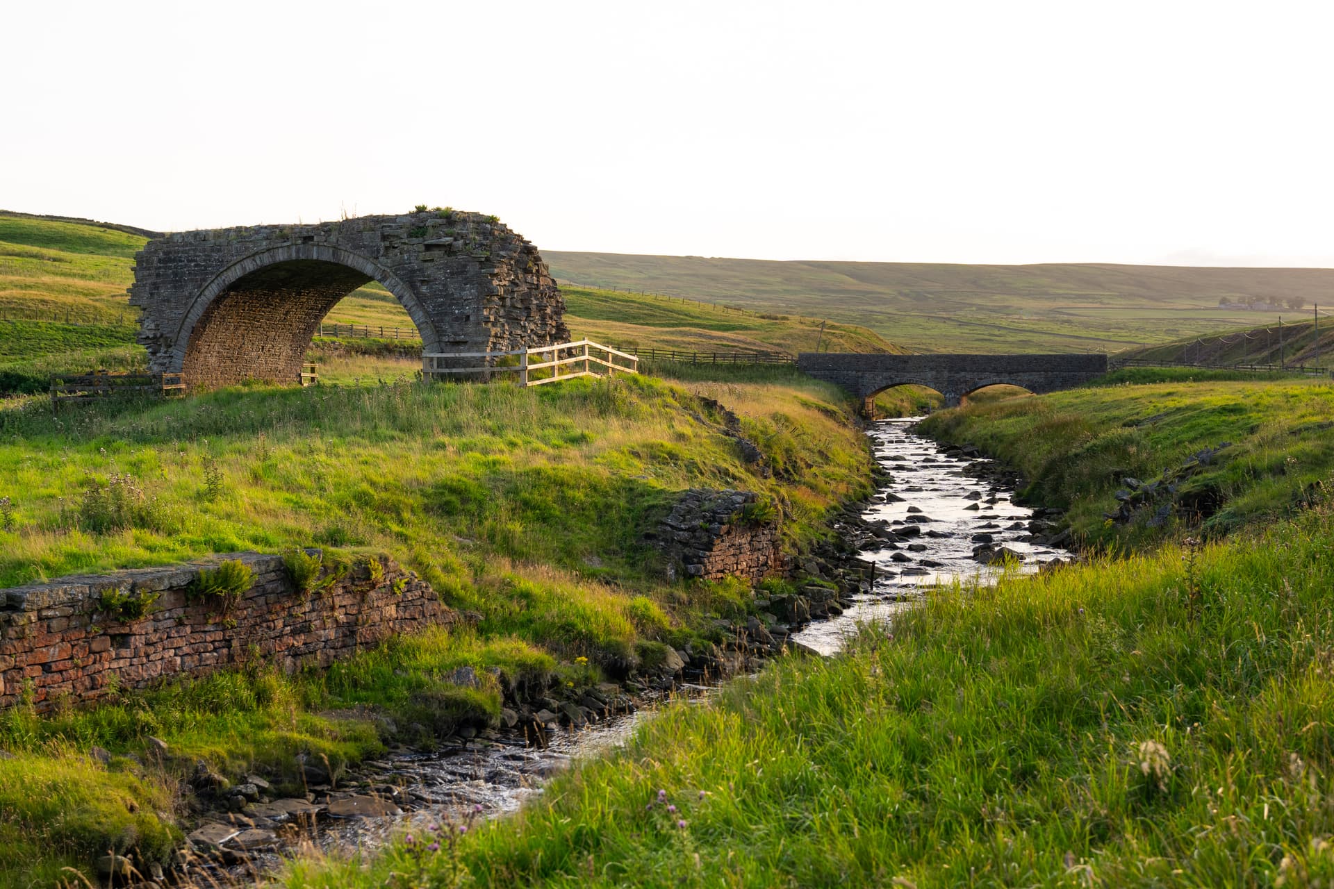 Stone arch bridge ruins over a rocky stream in a grassy valley, Rookhope Valley.