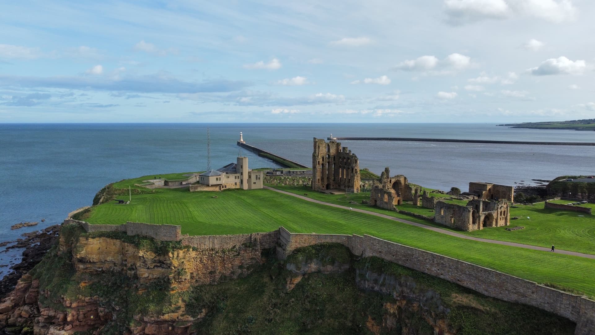 Tynemouth Priory ruins on grassy cliff overlooking the sea with a distant lighthouse.