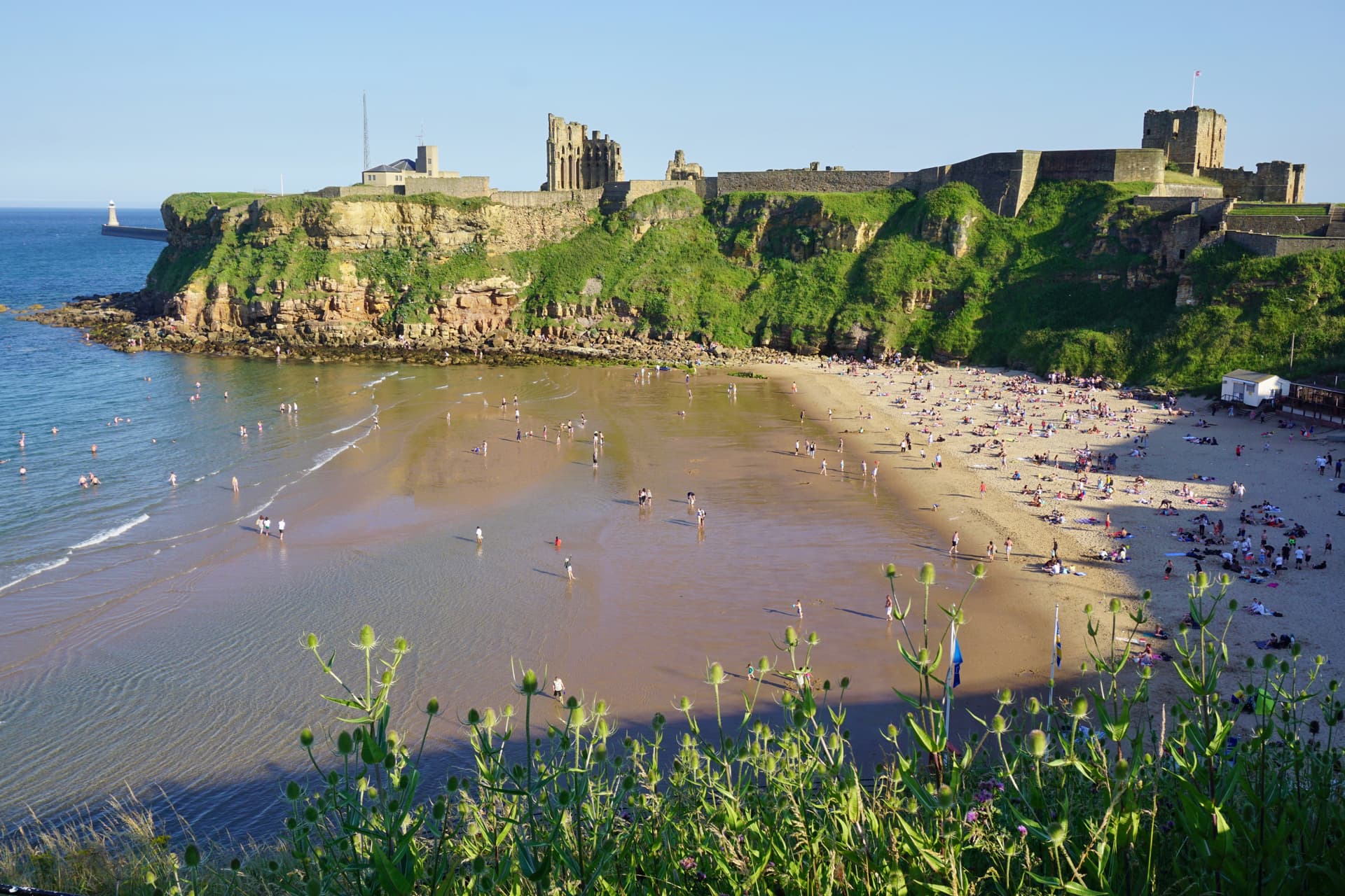 Beachgoers at Tynemouth with castle ruins atop a grassy cliff and a lighthouse.