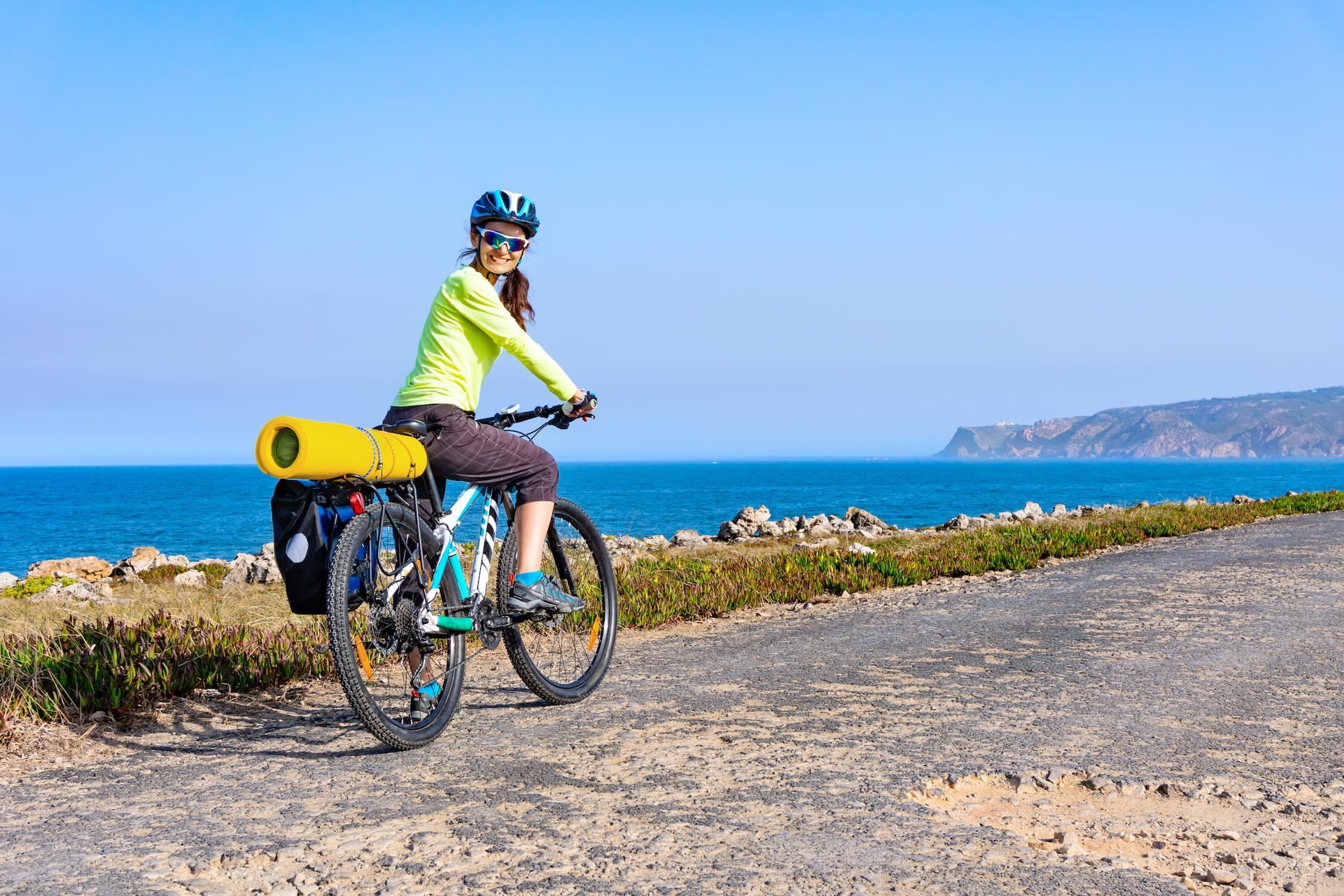 Cyclist with yellow mat touring on rough coastal road overlooking blue sea and distant cliffs.