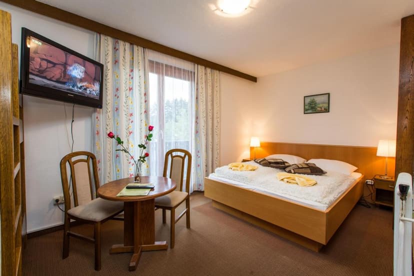 Hotel room with wooden bed, small dining table, chairs, and wall-mounted TV near balcony.