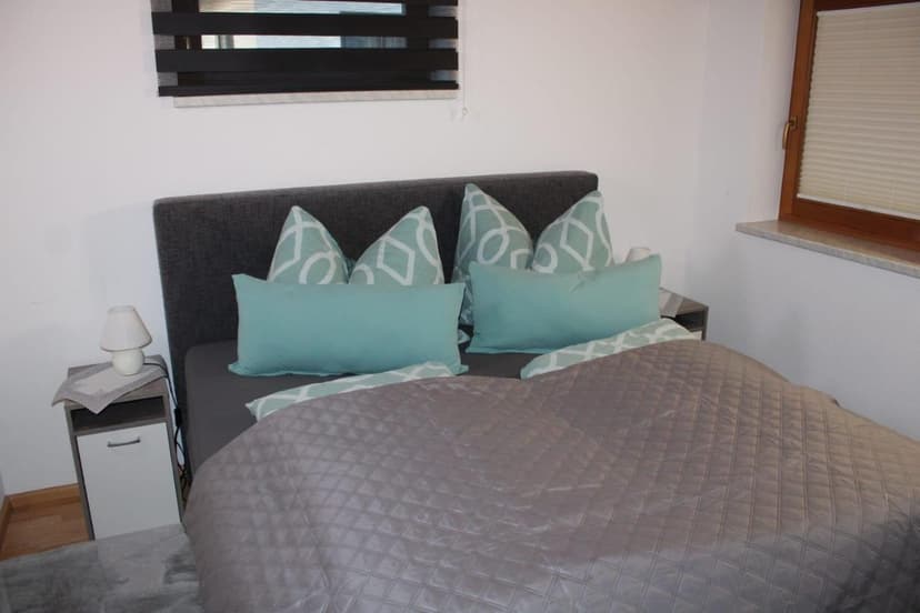 Bedroom with gray quilted bedspread, teal pillows, and nightstand with lamp in Ferienwohnung Waldheim.
