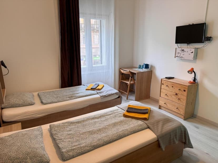 Twin beds in a simple room with a window, desk, and wall-mounted TV at Omega Guesthouse Budapest.
