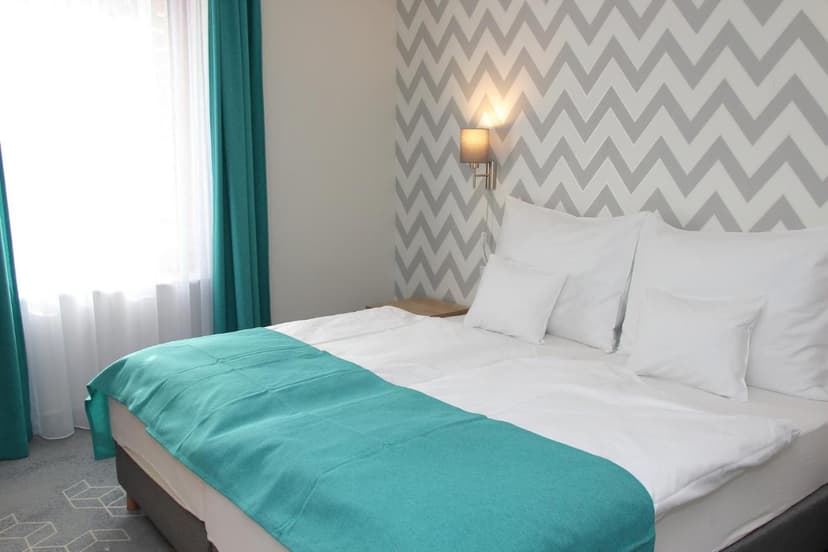 Hotel room with white bedding, teal throw, and gray chevron wallpaper near bright window.