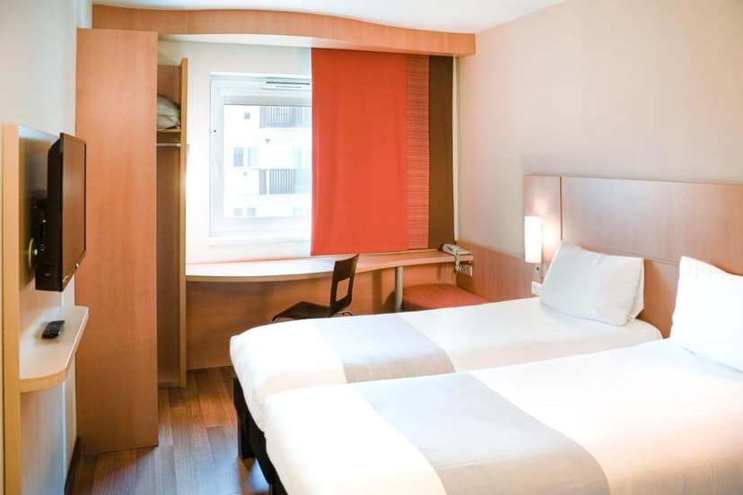 Twin beds in a hotel room with a desk, wall-mounted TV, and window with orange blind.