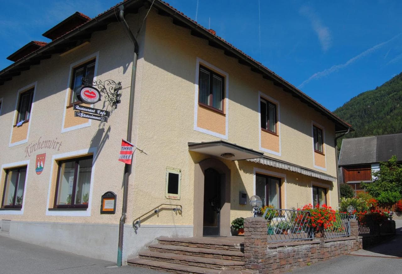 Pension Kirchenwirt building with "Restaurant Cafe Gästezimmer" sign near green mountain.